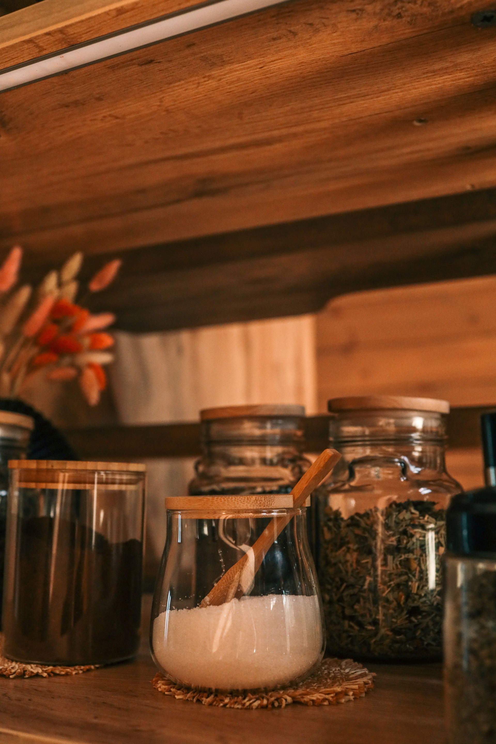 Close-up of a wooden kitchen shelf with glass jars filled with coffee, sugar, tea, and spices. Natural light highlights the warm tones of the cozy rustic interior